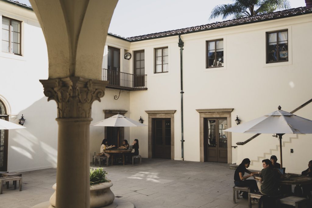Multiple groups of students engage in conversation within an outdoor courtyard, seated at tables shaded by umbrellas. To access this small courtyard lined with beautiful architecture, you should proceed through the hallway upon entering Hershey Hall. Photographed by Xiang Li/BruinLife.