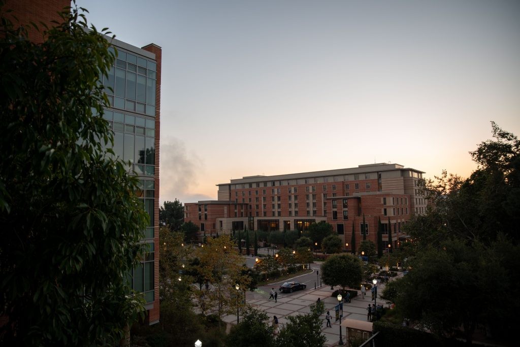 The sun sets behind the Luskin Conference Center, and the trees partially conceal the glass and brick of Engineering VI in the foreground. Accessible from the Bruin Reflection Space or from stairs directly below, the southwest corner of Ackerman Union overlooks the turnaround in Westwood Plaza. Photographed by Julia Gu/BruinLife.