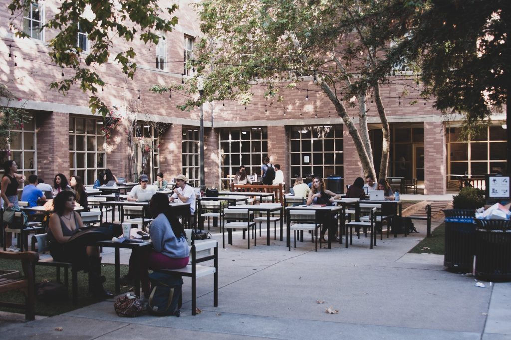 The courtyard of the UCLA School of Law building is often a quiet place to sit, with chairs and tables scattered around the area. High-reaching trees provide speckled shade and a serene atmosphere.