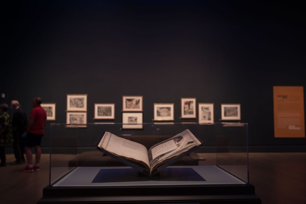 A large book stands in the room of the William Blake exhibit, with a wall of frames behind it. Blake was a poet, painter and printmaker, and he often combined the different mediums in his works. Photo by Katelyn Michel/BruinLife.
