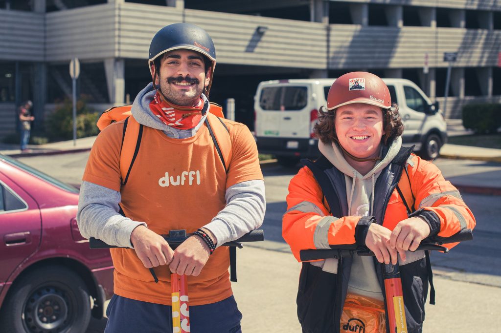 Aiden Seidle (right) and co-worker share a laugh while taking a break from scootering, decked out in vibrant orange safety vests and helmets, embodying the joy of a sunny day at work. Their camaraderie is as conspicuous as their safety gear, highlighting the lighter side of labor. Photographed by Olivia Maizes/BruinLife.