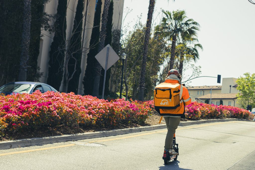 A Duffl employee, clad in the company's distinctive orange backpack, scoots along the Weyburn Graduate student apartments as the vibrant hues of spring blooms line the path of their delivery route. Photographed by Eliza Loventhal/BruinLife.