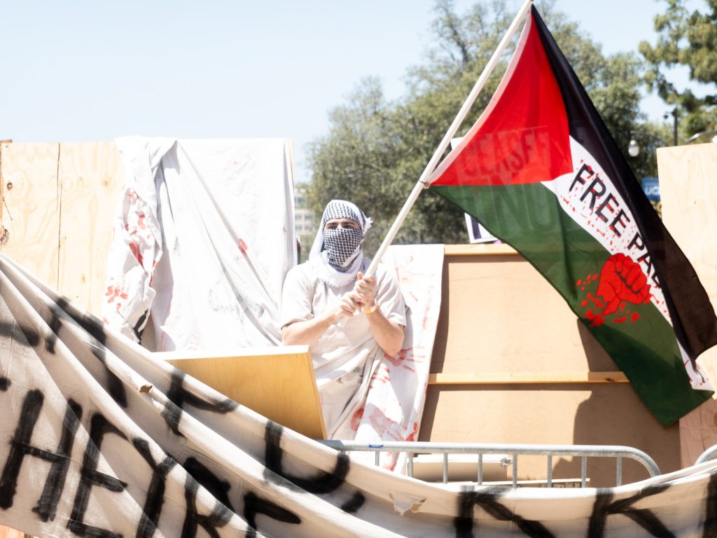A pro-Palestine protester waves a Free Palestine flag above the walls of the encampment. Student protesters constructed the barriers using pallet boards and fences, restricting access to a few controlled points. Photographed by Finn Martin/BruinLife.