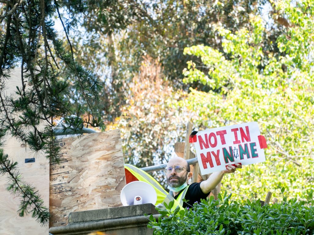 A pro-Palestine organizer appears from behind the encampment walls holding up a sign that reads "NOT IN MY NAME," with the "A" being written with the Star of David. The encampment spread overnight past the Shapiro Fountain, barricading Janss/Kuruvungna Steps and adjacent walkways.  Photographed by Finn Martin/Bruinlife.