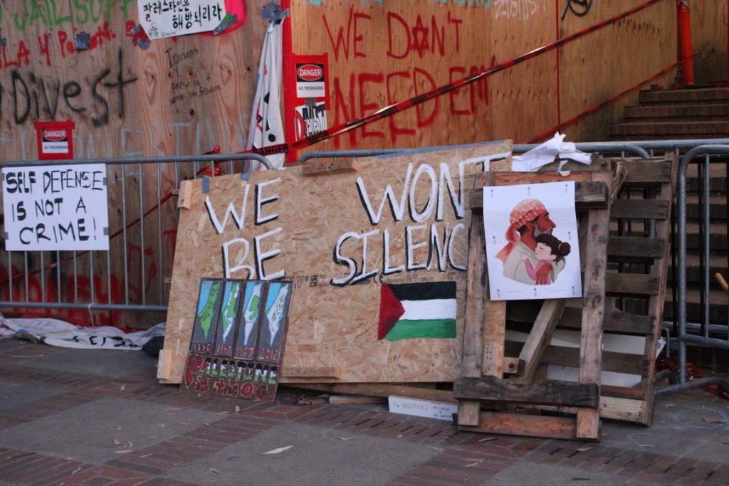 Pallet boards reading "SELF DEFENSE IS NOT A CRIME!" and "WE WON&squot;T BE SILENCED" rest against a barrier fence. Photograph provided by the Encampment Document Team.