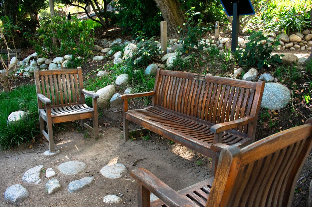 A wooden bench and two wooden chairs sit next to walkways lined with rocks. Seating can be found all throughout the Botanical Garden, such as these cute wooden benches.