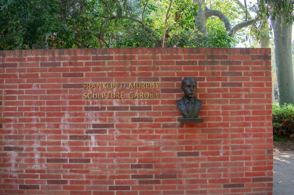 The Franklin D. Murphy Sculpture Garden entrance sign hangs on a brick wall. The sign includes a face sculpture of Franklin Murphy that protrudes out of the wall.