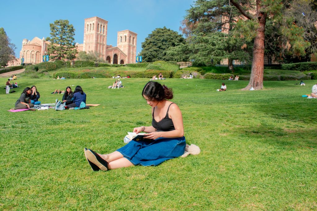 Sonja Cayetano, a fourth-year world arts & cultures student, sits on the grass along Janss/Kuruvungna Steps and reads a book. Royce Hall stands in the background.