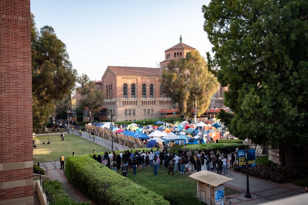 A view of the Palestine solidarity encampment and the wooden barricade created between the protesters and the rest of Dickson Plaza. A crowd of students observe the scene standing between Royce Hall and Haines Hall. Photographed by Finn Martin/BruinLife.