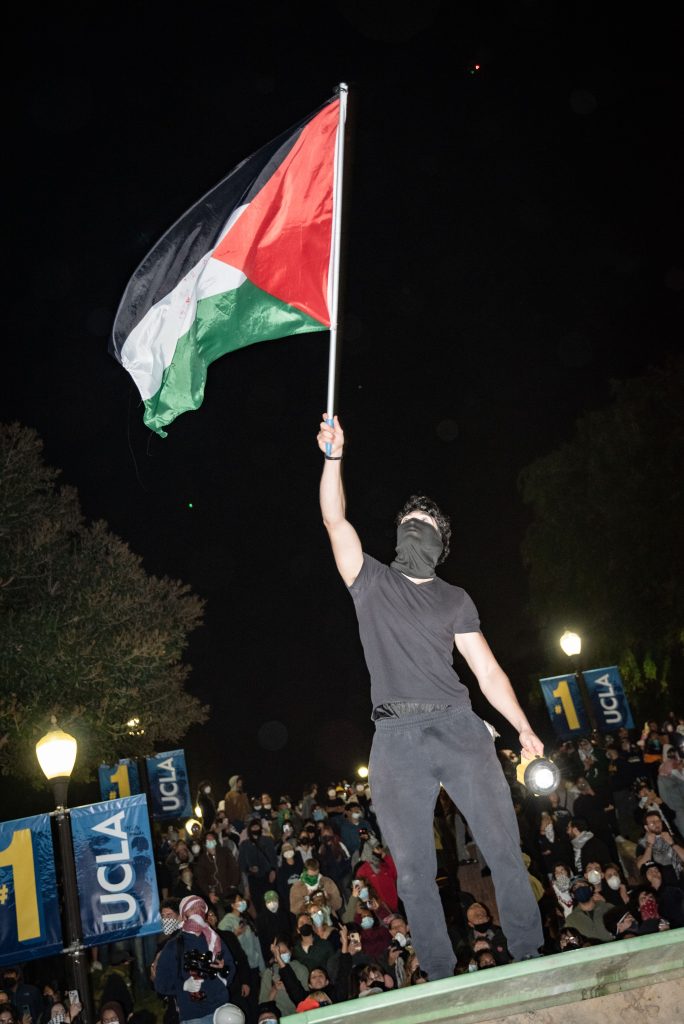 A pro-Palestine protester waves a banner as a show of triumph while standing atop dumpsters that were moved to the bottom of Janss/Kuruvungna Steps. Protesters from inside the encampment expelled the police from their hold on Janss/Kuruvungna Steps toward the field of Wilson Plaza. Photographed by Finn Martin/BruinLife.