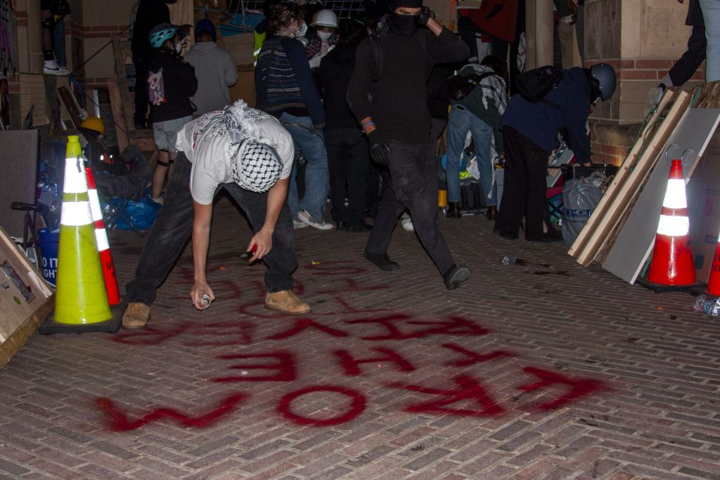 A solidarity protester uses red spray paint to write "FROM THE RIVER TO THE SEA" on the grounds of Royce Hall inside the encampment. Photographed by Emily Chandler/BruinLife.