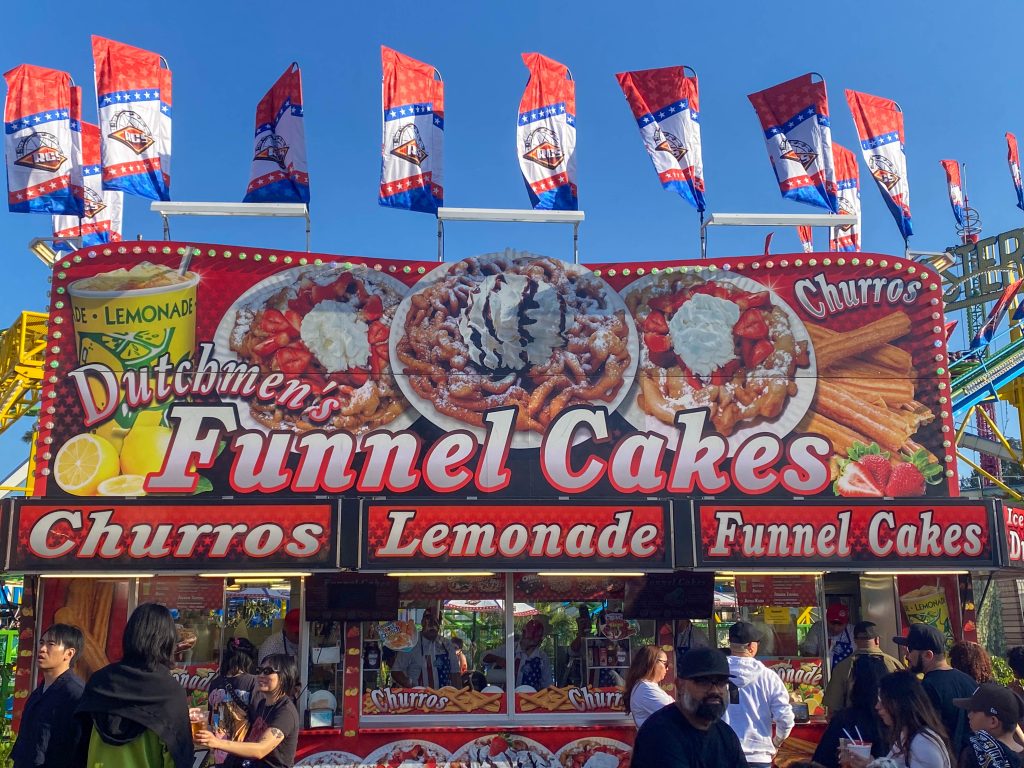 A food stall advertising funnel cakes, churros and lemonade tends to a line of visitors in need of a quick snack or refreshment from the heat.