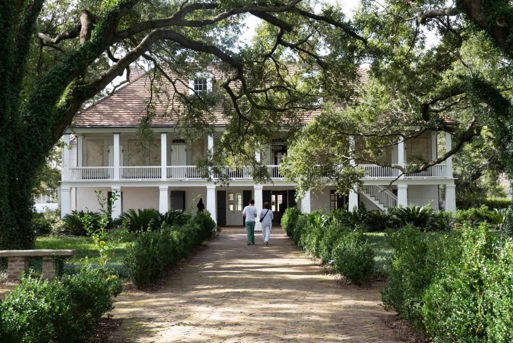 The garden facing facade of the enslavers plantation manor.  Ivy covered oak trees enclose the promenade leading to the three story home, furnished with a false façade of painted marble. Photographed by Finn Martin/BruinLife.