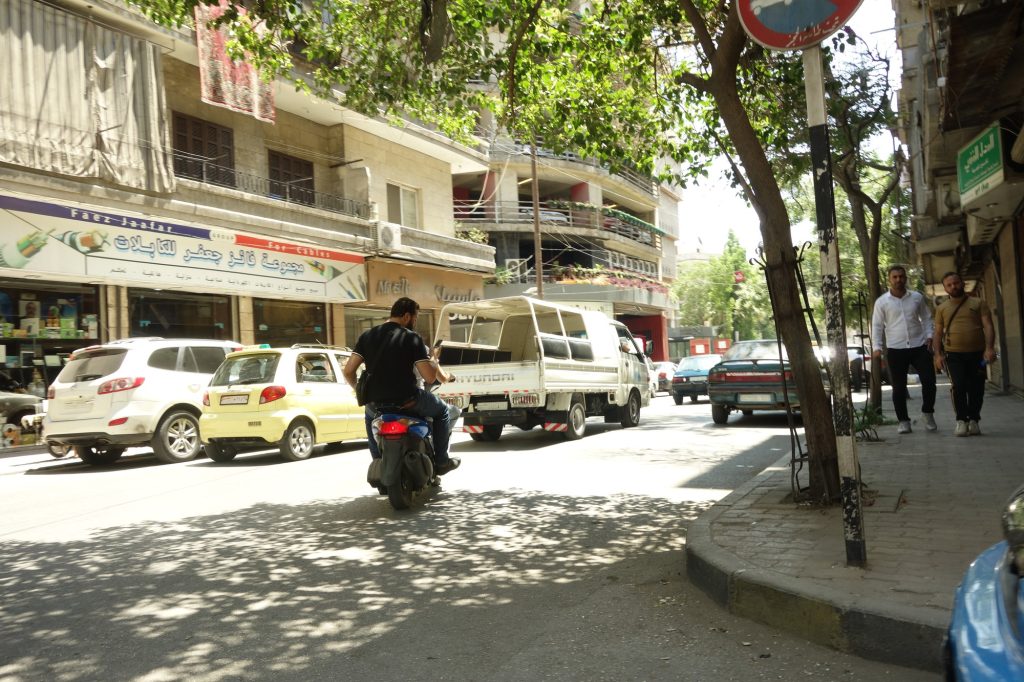 A motorcyclist rides through traffic while pedestrians walk along the sidewalk on a vibrant street in Syria in July 2022. The photo was taken by Celia during her first visit in 12 years since the start of the civil war in 2011.