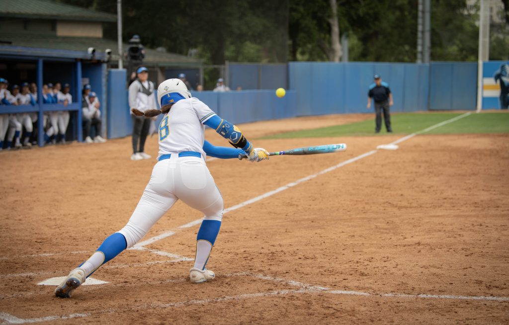 Jessica Clements sends the ball down the left field as her teammates and coach watch from the dugout. Photographed by Sapna Drew/BruinLife.